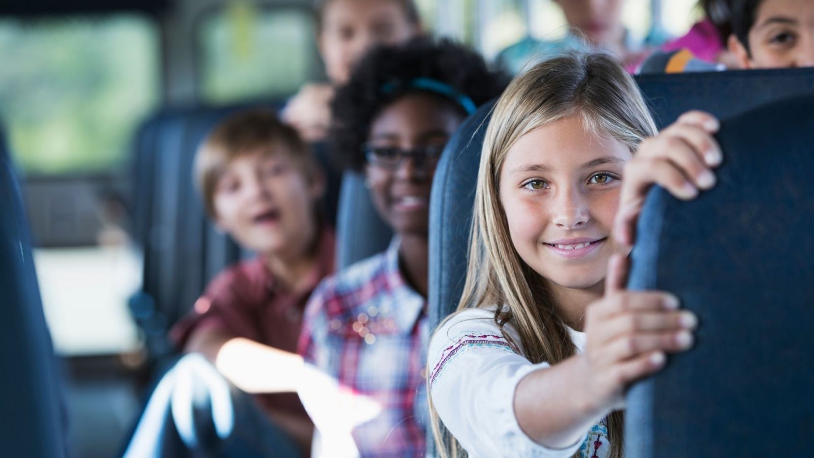A young girl sitting on a bus with her hand on the seat in front and looking down the aisle of the bus